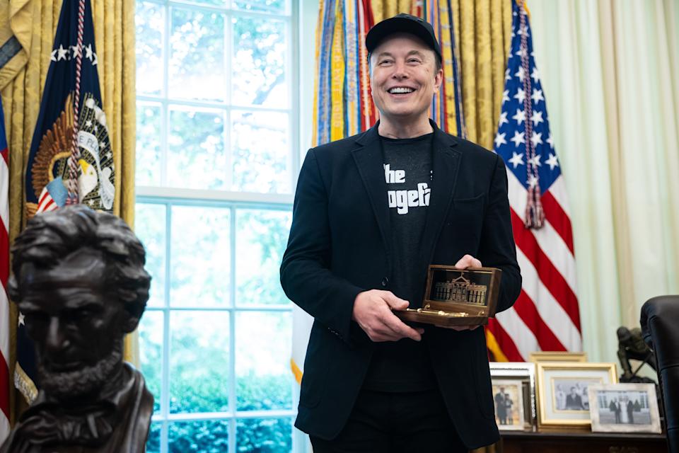 A person smiling in an office, holding a model building, with flags in the background and a bust of a historical figure nearby