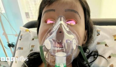 A manikin doll wearing an oxygen mask with brown hair leans back against a pillow