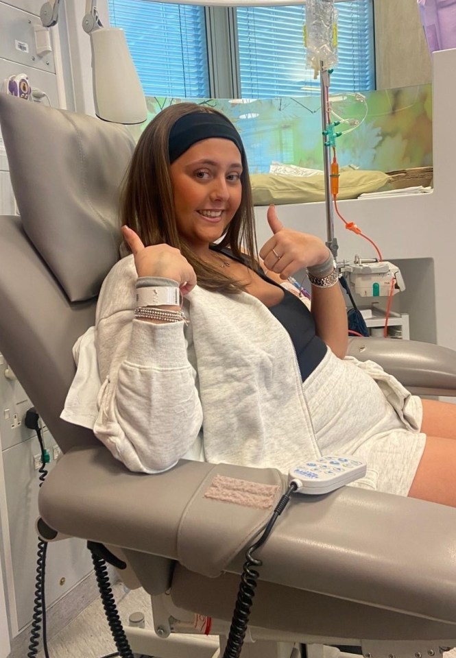 A young woman gives a thumbs-up while sitting in a hospital chair during an IV treatment.