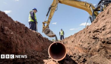 Two builders laying pipework on housing building site.  They are wearing blue overalls, blue hard hats and yellow high-visibility gilets.  They are standing over a trench as a digger empties soil on top of a plastic pipe.