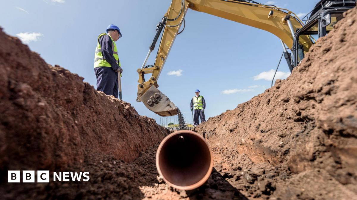 Two builders laying pipework on housing building site.  They are wearing blue overalls, blue hard hats and yellow high-visibility gilets.  They are standing over a trench as a digger empties soil on top of a plastic pipe.
