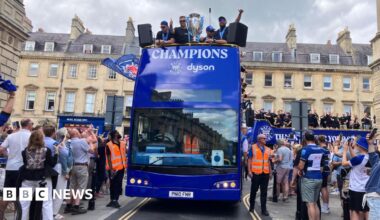 A blue double decker bus with Champions written on the front alongside Bath Rugby's logo. There are several Bath Rugby players on the top deck with a big trophy. There are crowds on either side of the road by the bus, many of whom are in Bath Rugby clothing.