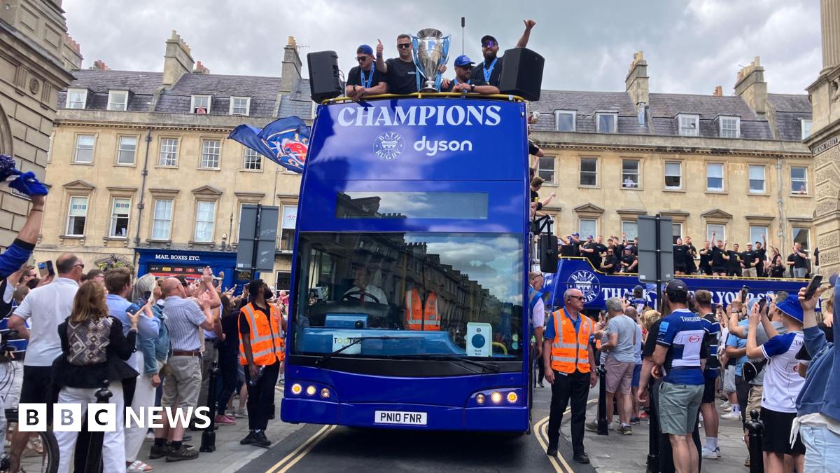 A blue double decker bus with Champions written on the front alongside Bath Rugby's logo. There are several Bath Rugby players on the top deck with a big trophy. There are crowds on either side of the road by the bus, many of whom are in Bath Rugby clothing.