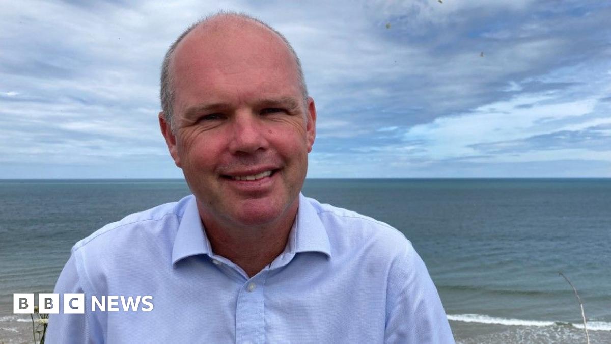 Andrew Raine, a man standing outside at Sheringham beach. He is wearing a blue shirt and is looking directly at the camera and smiling.