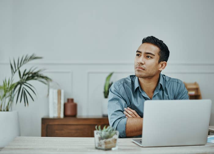 A man sits at a desk with a laptop, looking thoughtfully to the side