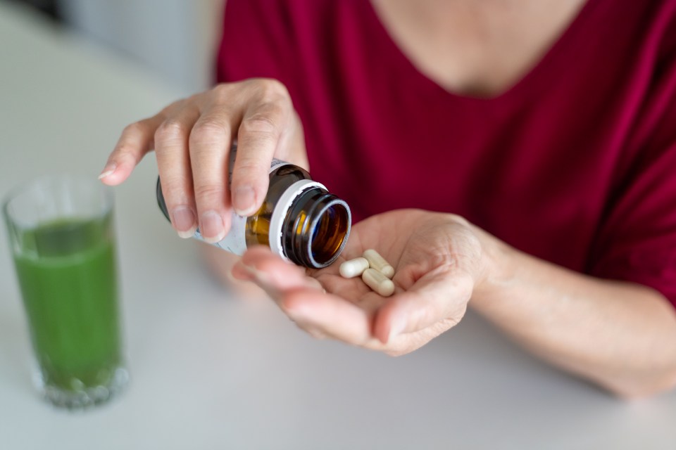 Close-up of hands pouring vitamins from a bottle into a hand; a green smoothie is in the background.