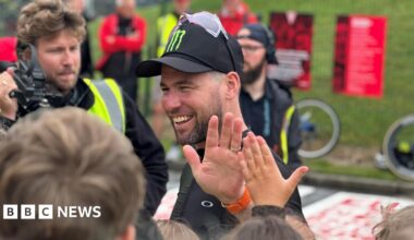Sir Mark Cavendish, wearing a black cap with sun glasses on top of it, giving a high five to a young fan while a man with a film camera looks on.