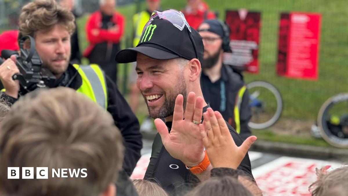 Sir Mark Cavendish, wearing a black cap with sun glasses on top of it, giving a high five to a young fan while a man with a film camera looks on.