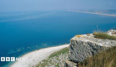 A view over Lyme Bay from Westcliffs in Portland. The weather is sunny. There is a stone in the foreground at the top of a cliff. The cliff is visible below as well as a beach and vast expanse of deep blue sea.