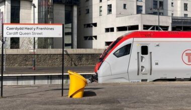 A Transport for Wales train pulled into Cardiff Queen Street, with the red T logo in a circle displayed on the side of the grey and red train. There is a gritting bin in shot, and a sign for Cardiff Queen Street, and its Welsh version Caerdydd Heol y Frenhines, at the left hand side.