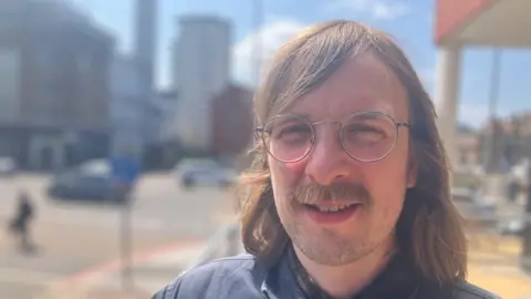 Head and shoulders of a man with long hair, a moustache and large thin rimmed glasses stood outside
