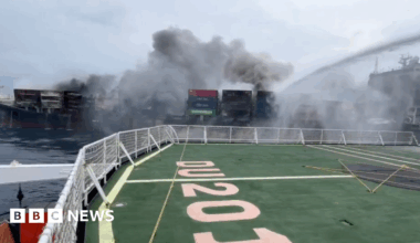 A ship is on fire. Thick dark smoke rises from the containers on its deck. In the foreground, we see the deck of another vessel with railings and a large green area marked “0205.” A strong stream of water sprays from the boat, aiming at the fire.