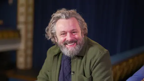 BBC Michael Sheen, who has long grey hair and a grey beard and is wearing a navy jumper and green jacket as he smiles at the camera. 
