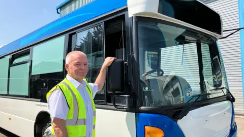 Henry Godfrey-Evans/BBC Bill Hiron in a high-visibility vest with his hand on the wing mirror of his bus