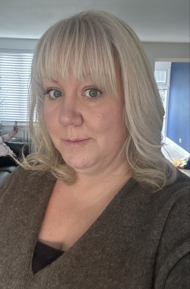 Close-up selfie of a woman with blonde hair and bangs, wearing a brown sweater.