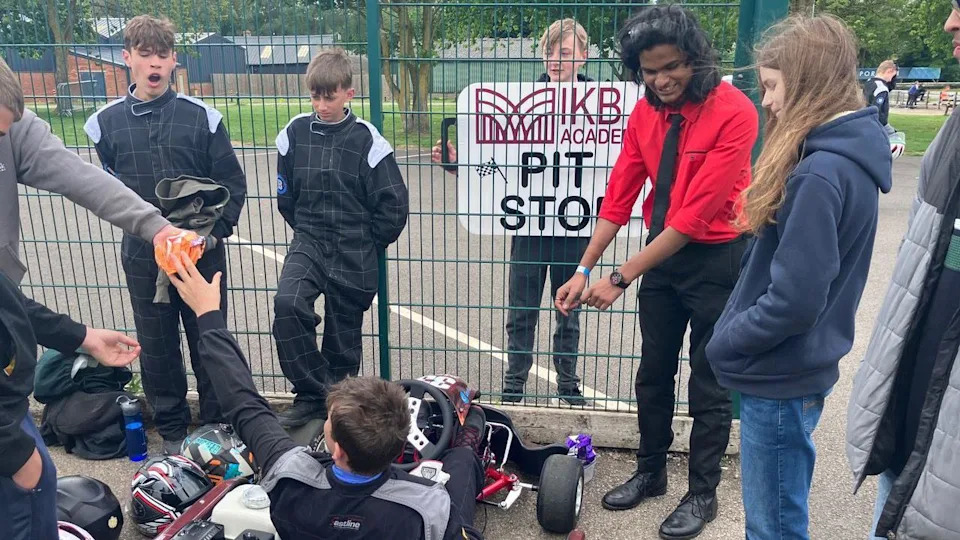 A group of young people in motorsport padded overalls stand over another young person in a small motorised go-kart