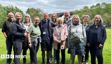 A group of walkers are pictured in a field in Reddish Valley Park, Stockport. They are grouped closely together and are all smiling on a sunny day.