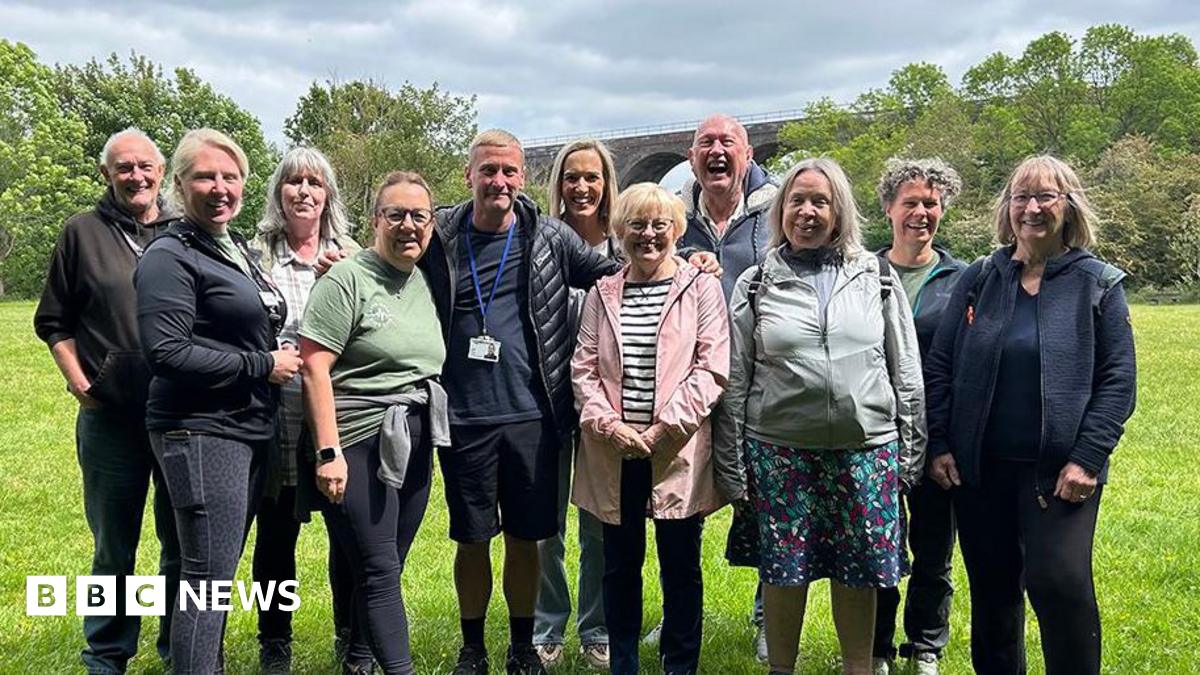 A group of walkers are pictured in a field in Reddish Valley Park, Stockport. They are grouped closely together and are all smiling on a sunny day.