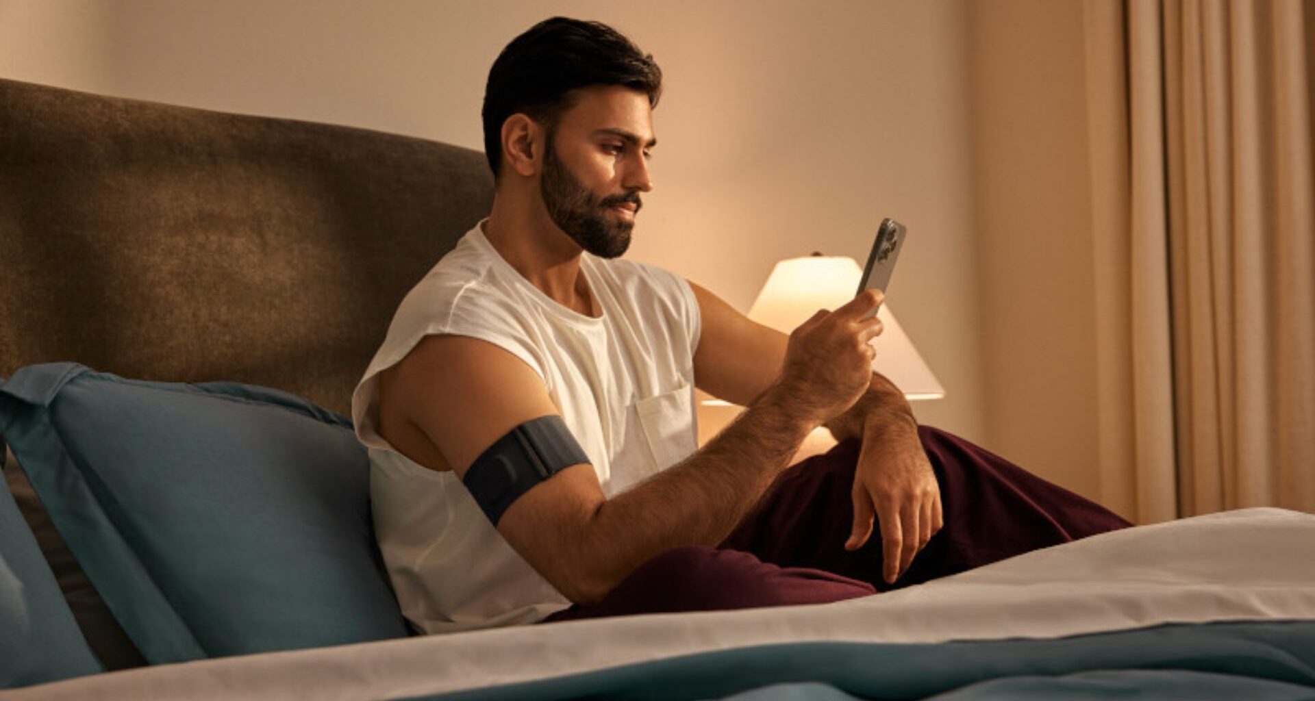A man in white vest sitting up in bed checking phone with Garmin Index Sleep Monitor on right arm