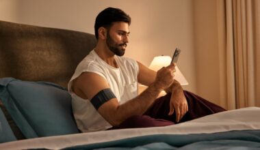 A man in white vest sitting up in bed checking phone with Garmin Index Sleep Monitor on right arm