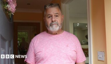 Paul Thomas wearing a pink t-shirt is standing in his doorway and looking straight at the camera.