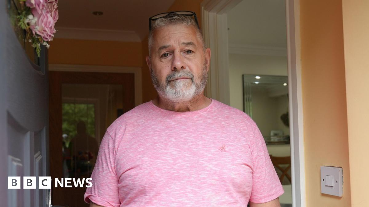 Paul Thomas wearing a pink t-shirt is standing in his doorway and looking straight at the camera.