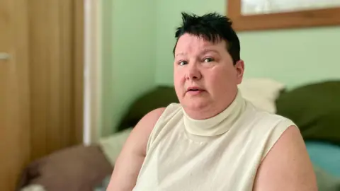 Mark Norman BBC A woman sitting on a sofa wearing a white sleeveless top.