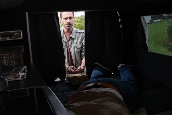 John holding a syringe as he stands by unconscious Robert in Emmerdale