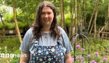 Elinor Casey, with long curly brown hair, a grey top and flowery dungarees stands in front of a garden with trees and purple flowers to the right of the picture.