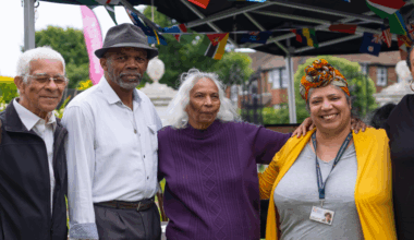 Two men and three women, including Shirley Williams, pose for a photograph at a Windrush event from 2024. They stand under a gazebo draped in flags from a variety of nations.