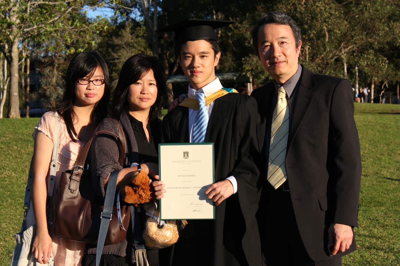 A young man in academic regalia, holding a degree, poses with his family.