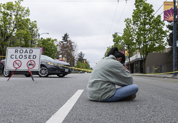 A woman sits on the pavement praying with a 'road closed' sign nearby