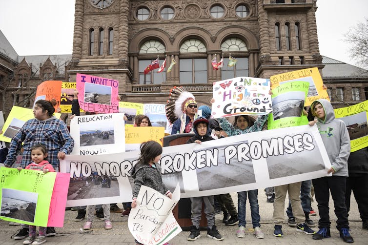 people at a protest with a large banner that reads: no more broken promises. A child carries a poster reading: we need higher ground