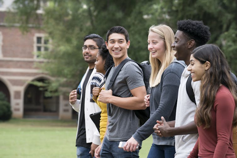 A group of six smiling students walks outside on a college campus.