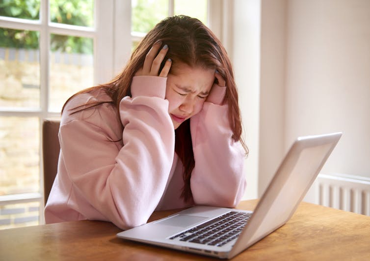 A student with dark hair sits in front of a laptop computer crying with her head in her hands.