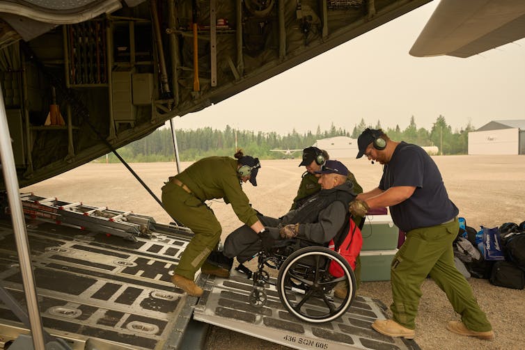 Men in green military fatigues help a man in a wheelchair up a plane ramp