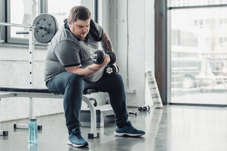 An overweight man with tattoos on his arms performs a barbell curl at the gym while seated on a bench.