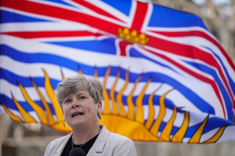 A woman with short dark blond hair in front of a B.C. flag.