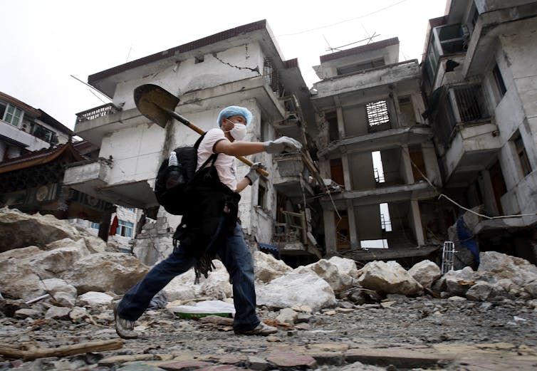 A volunteer carrying a shovel over his shoulder walks past boulders and a severely damaged building.