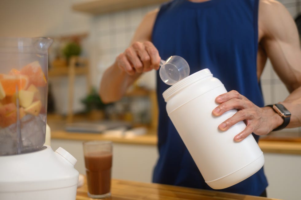 man adding a scoop of chocolate protein powder into a blender filled with chopped fruits, preparing nutritious shake in the kitchen.