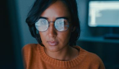 Woman in glasses reflecting code, wearing a rust-coloured jumper, working at a computer screen