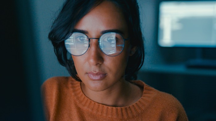 Woman in glasses reflecting code, wearing a rust-coloured jumper, working at a computer screen