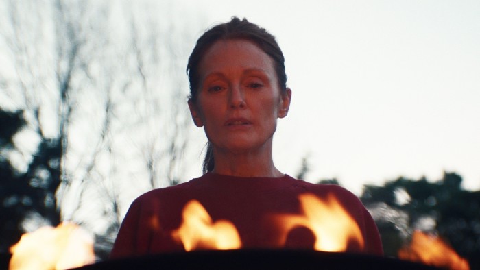 A woman stands facing us with flames in front of her