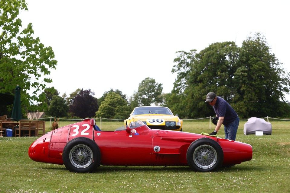 A red vintage race car being polished on a lawn, with a yellow sports car in the background.