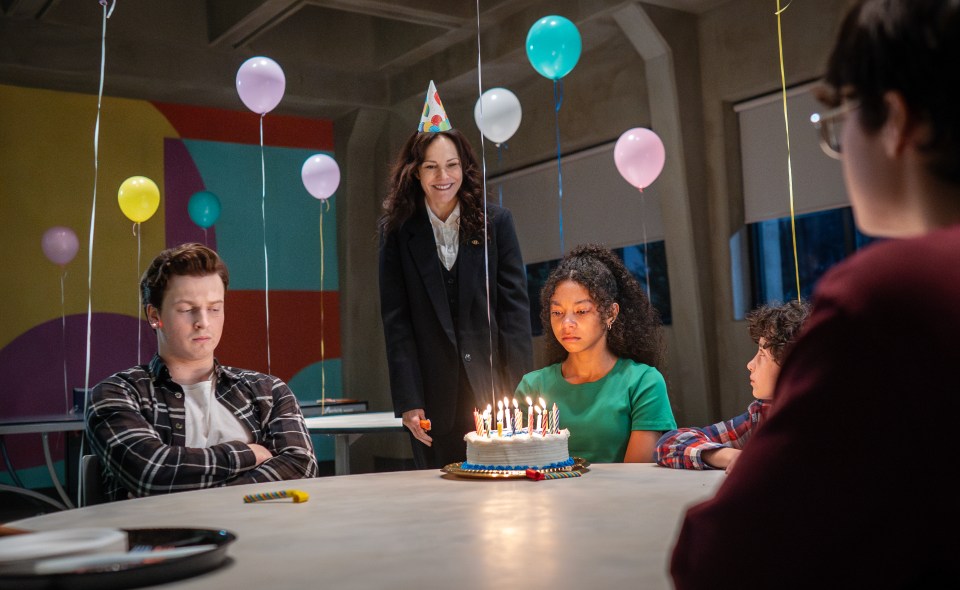 A birthday cake with lit candles is at the center of a table, surrounded by a group of young people and an adult woman wearing a party hat.