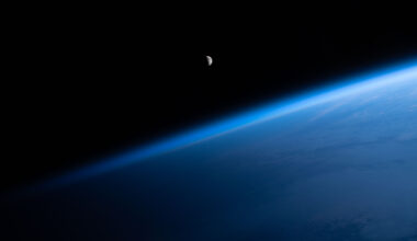 The Moon is a small gray circle with less than half of it visible. Below it, Earth's atmosphere is a brilliant blue haze making a diagonal line across the image.