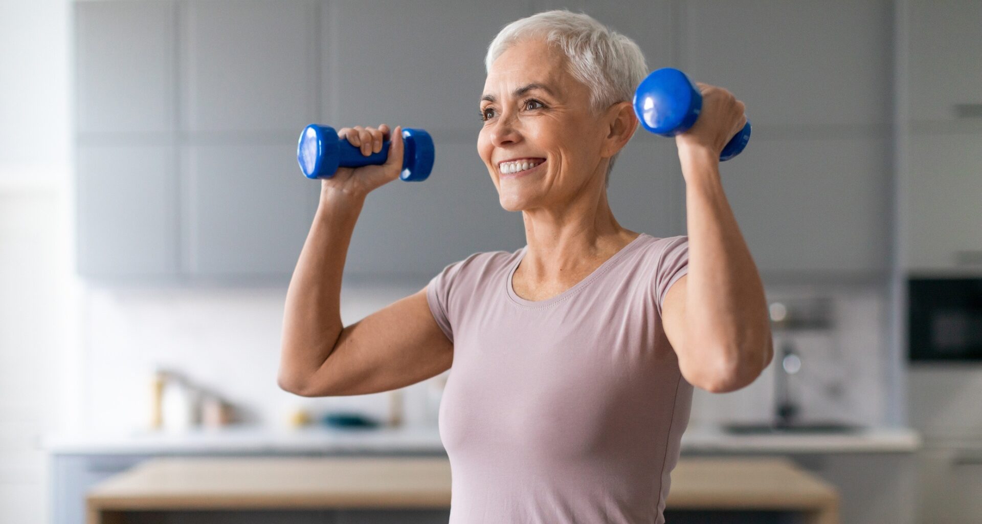 woman from waist up, holding light blue dumbbells in each hand ready to do an overhead press, smiling. she's wearing a light pink tshirt in a kitchen setting that's blurred in the background.