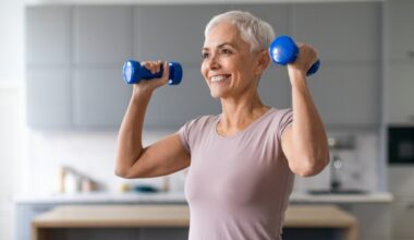 woman from waist up, holding light blue dumbbells in each hand ready to do an overhead press, smiling. she's wearing a light pink tshirt in a kitchen setting that's blurred in the background.