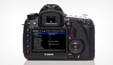 A Canon DSLR camera viewed from the back, displaying an on-screen settings menu labeled "Overlay" with various options such as Zebras, Focus Peak, and Histogram. Several buttons and dials are visible around the screen.