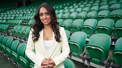 Isa Guha wears a cream coloured suit and stands in front of green stadium seats